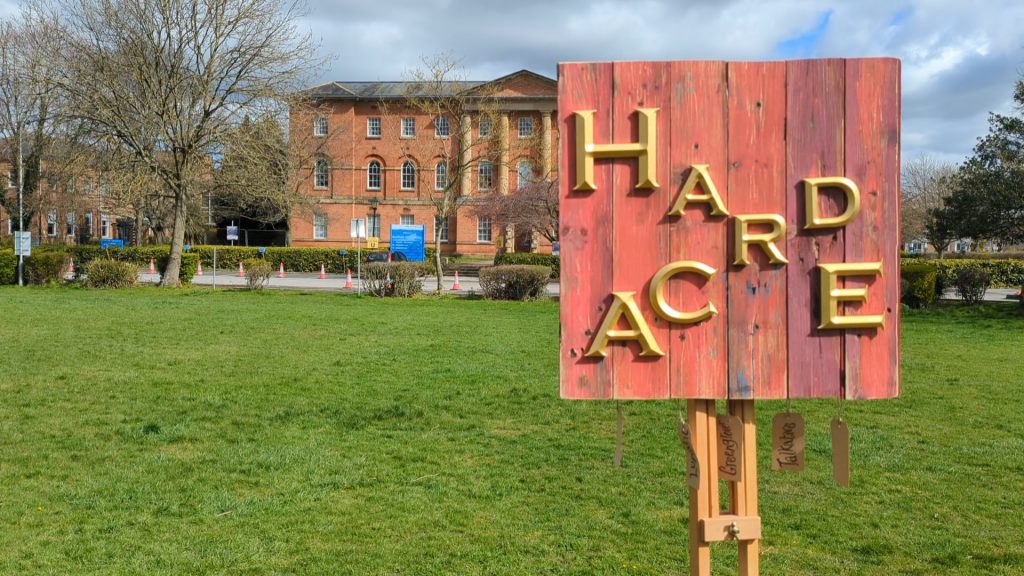 Photo of a piece of art on an easel in front of Bootham Hospital York. Made of reclaimed materials from The Groves, York, including part of an old backyard door, and signage letters from, the now closed, Castle Howard Ox pub on Townend Street, 65 x 65 cm, Stephen Lee Hodgkins, 2023.
