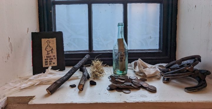 Image of a window sill, with from left to right, a handprinted booklet that reads ‘I thought you were going to look over that job’ and a print of a goose, a pile of feathers, coins, burnt wood, some hay, several horseshoes, a piece of cloth ripped from an apron and a bottle with a note in it.